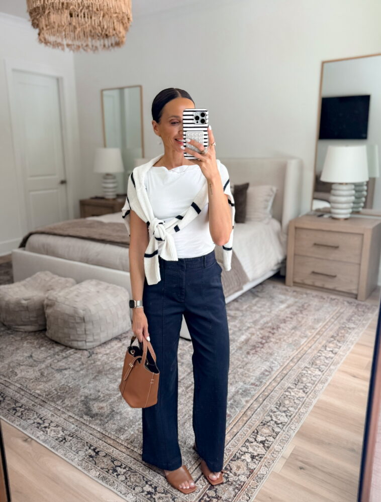 Classic workwear outfit with white t-shirt, striped sweater layered over shoulders, navy wide-leg trousers, brown tote bag, and heeled sandals.